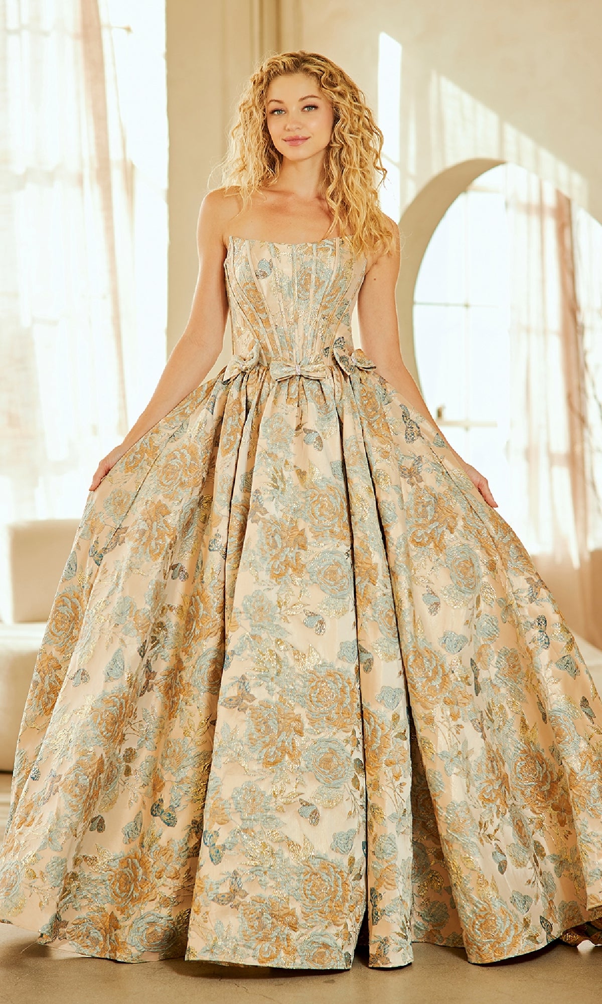 A woman stands indoors wearing the Amelia Couture 8068 Lace-Up Print Prom Ball Gown—a strapless, long floral dress in pale blue and gold—gracefully holding the sides of her gown.
